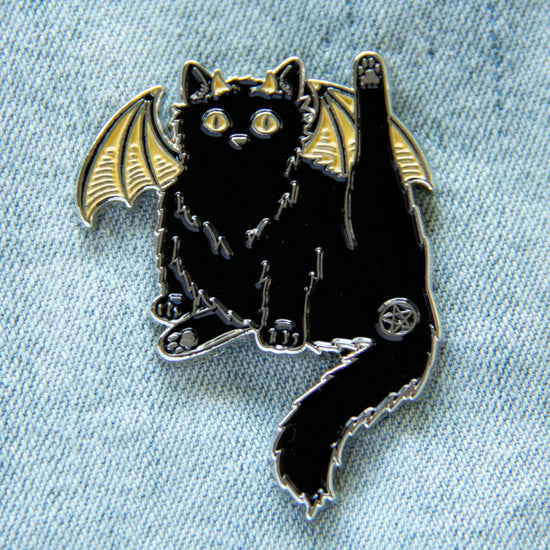 An enamel pin in the shape of a black cat, with yellow bat-like wings, affixed to light blue denim. One of the cat’s hind legs is lifted, and the cat’s butthole is covered by a pentagram.