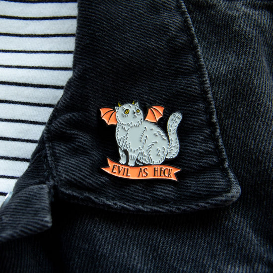 A grey cat with orange bat wings and yellow horns, attached to a dark denim collar. Under the cat is an orange ribbon with black text saying "Evil as heck."