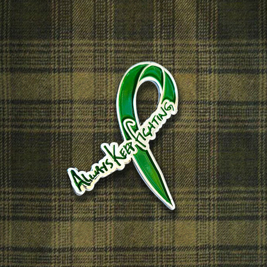 A green pin in the shape of a ribbon, with handwritten text across one end saying "always keep fighting." Behind the pin is a green plaid background.