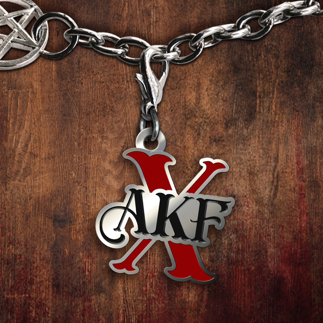 A metal charm on a chain, against a wooden background. The charm is shaped like a large red X, with the letters AKF on the front in black. In the top left corner is an anti-possession charm attached to the chain.