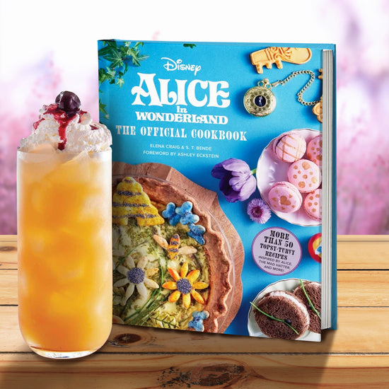 A blue book on a wood table, next to a glass filled with an orange beverage, topped with whipped cream and a cherry. On the cover is white text saying "Alice in wonderland, the official cookbook." Various cakes and cookies are shown on the cover, next to flowers and a pocketwatch. In the background are purple flowers.