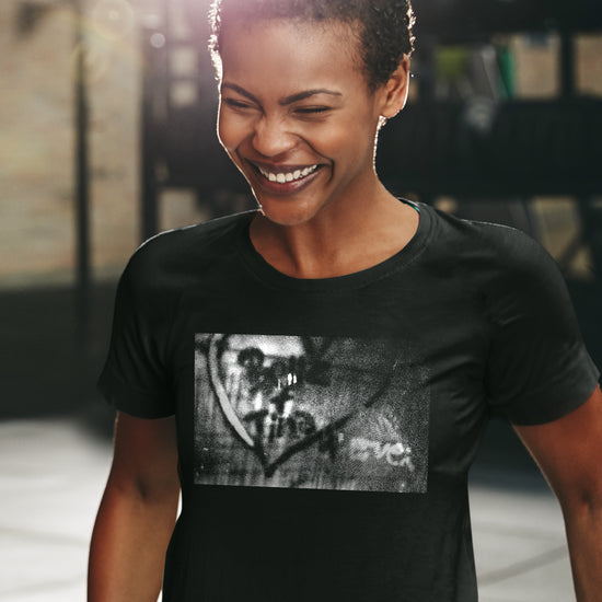 A female model wearing a black T-shirt, in front of a brick wall.. On the shirt is a black and white wall with Bette + Tina 4 Ever spray painted inside a heart.