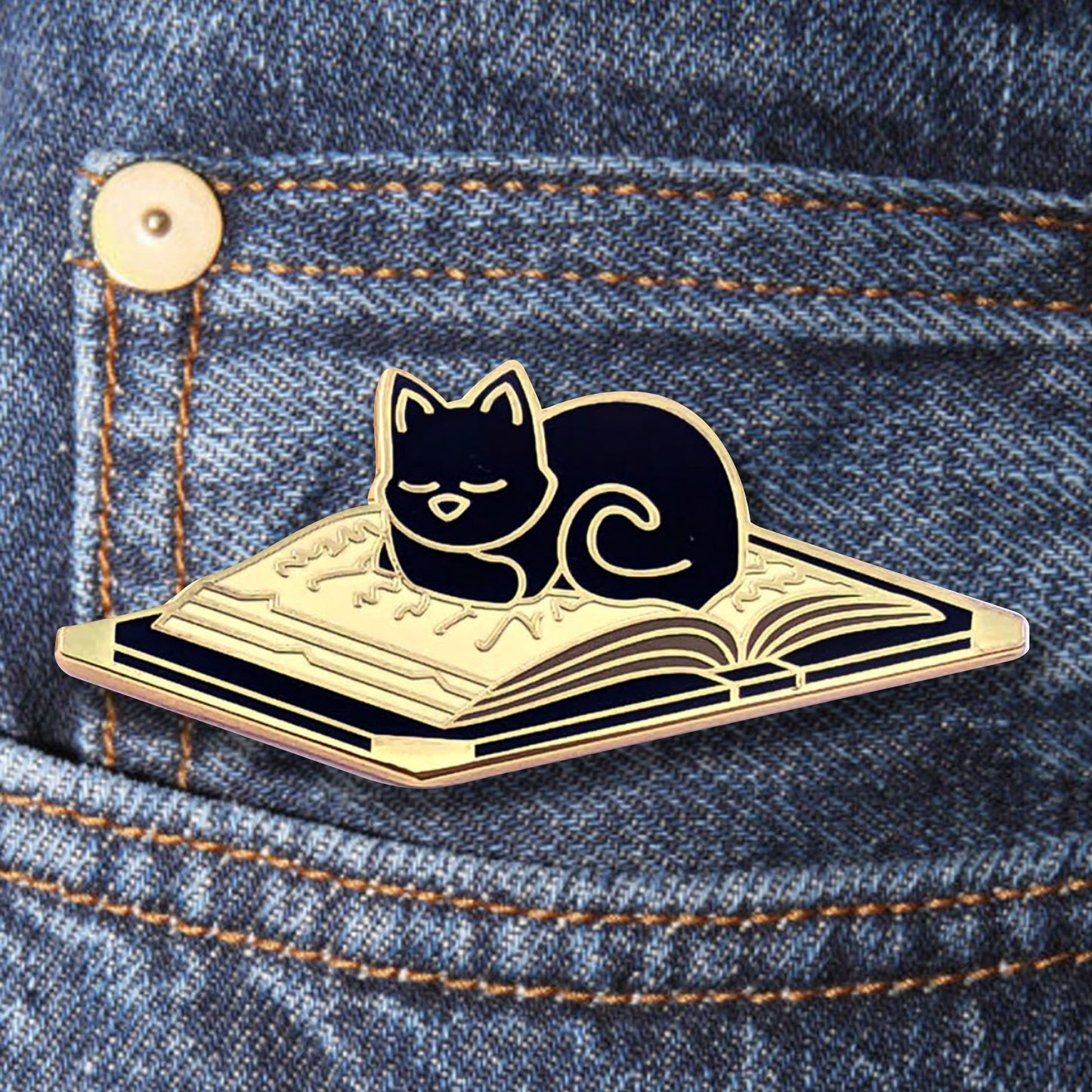 Close up of a gold colored enamel pin on a denim pocket. The pin is shaped like an open book with a black cat sleeping on the pages.
