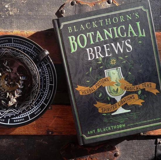 A black book on a brown wood table. A green and white border runs along the edges of the cover. At the top in green and white text is "blackthorn's botanical brews." In the center is a drawing of a white goblet with green liquid in in, with green vines surrounding it. At the sides of the goblet are yellow banners, with black text saying "herbal potions, magical teas, spirited libations." Next to the book is a black teacup and saucer, with white text and lines depicting zodiac signs.