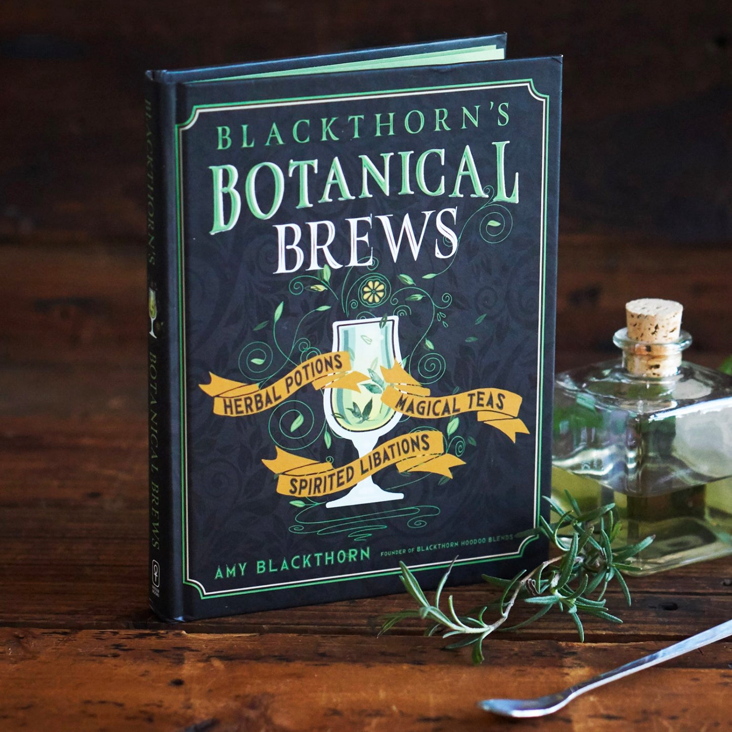 A black book on a wood table. A green and white border runs along the edges. At the top in green and white text is "blackthorn's botanical brews." In the center is a drawing of a white goblet with green liquid in in, with green vines surrounding it. At the sides of the goblet are yellow banners, with black text saying "herbal potions, magical teas, spirited libations." Next to the book is a glass vial, a sprig of herbs, and a metal cocktail stirrer.