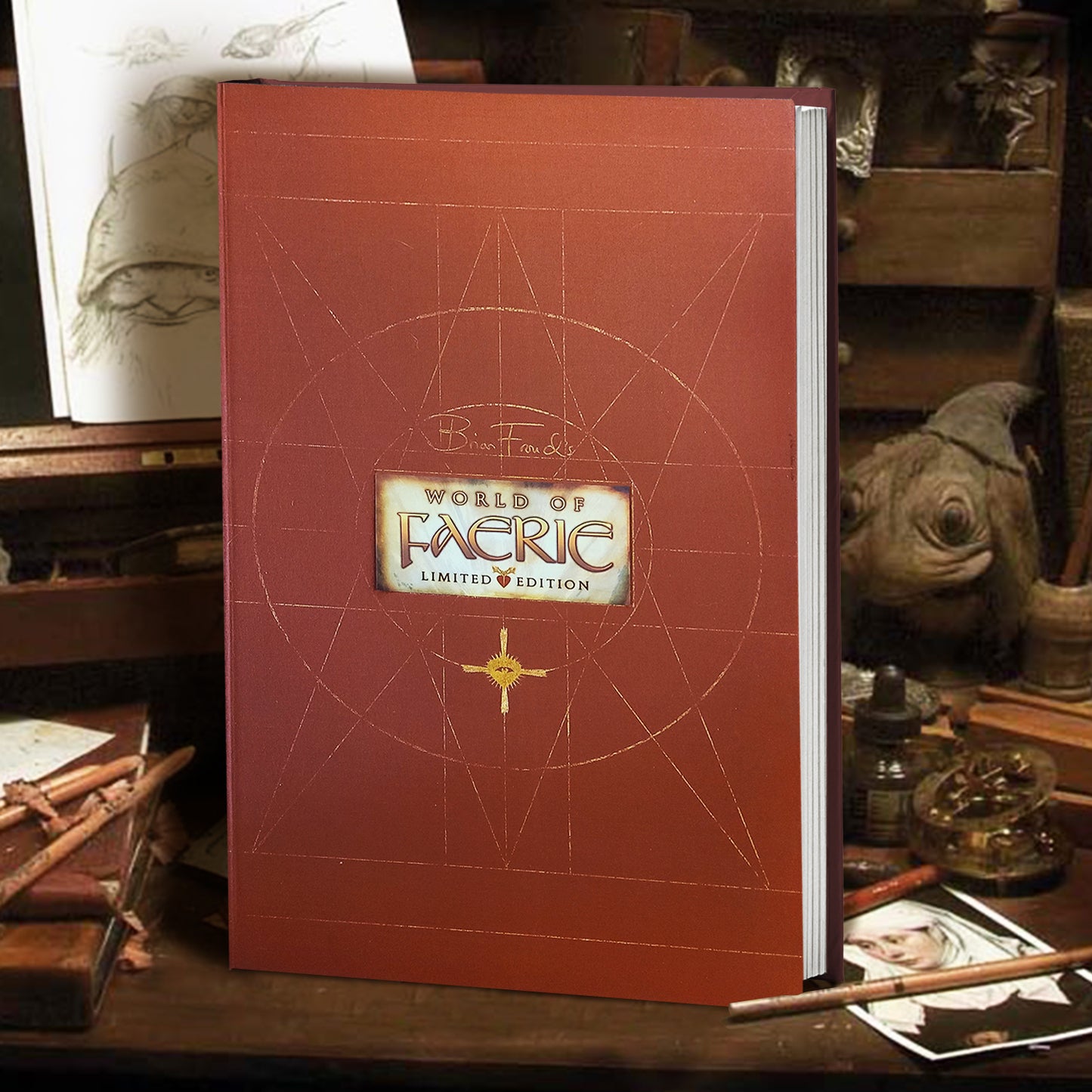 A brown hardcover book on a desk. At the center of the cover is a parchment-colored rectable with yellow text saying Brian Froud's World of Faeire, limited edition." Behind the book are old fashioned art tools, strwn about in small wooden drawers. A few drawings are scattered around the desk.