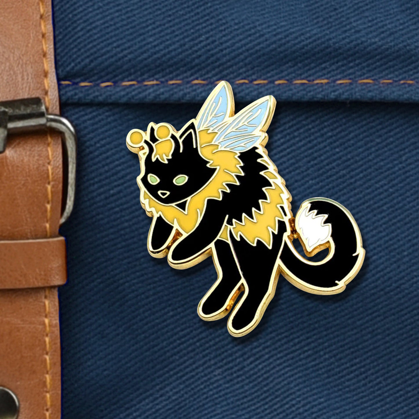 A black-and-yellow 'bumble cat' enamel pin. The pin is roughly in the shape of a fuzzy bumble bee, with a black cat head, tail, and four legs, as well as two small antennae and a small pair of bee wings. The pin is set against a navy canvas bag. 