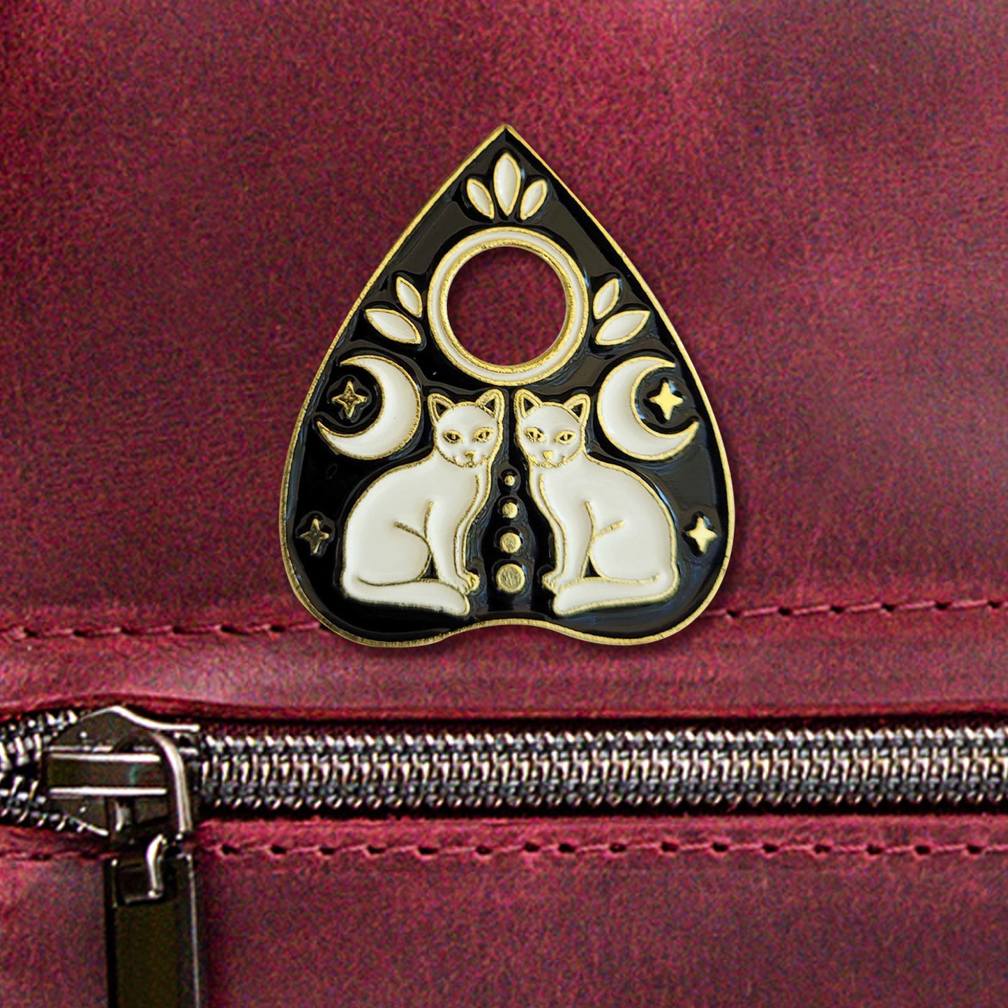 A close-up photo of a black-and-gold enamel pin in the shape of a planchette. The pin depicts two identical white cats facing each other, each with a moon and two small stars above/around them.