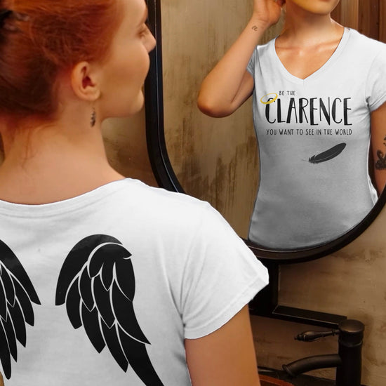 A female model looking in a mirror. She's wearing a white v-neck t-shirt that says "Be the Clarence you want to see in the world" with a halo and black feather on the front, and a black angel wing print on the back.