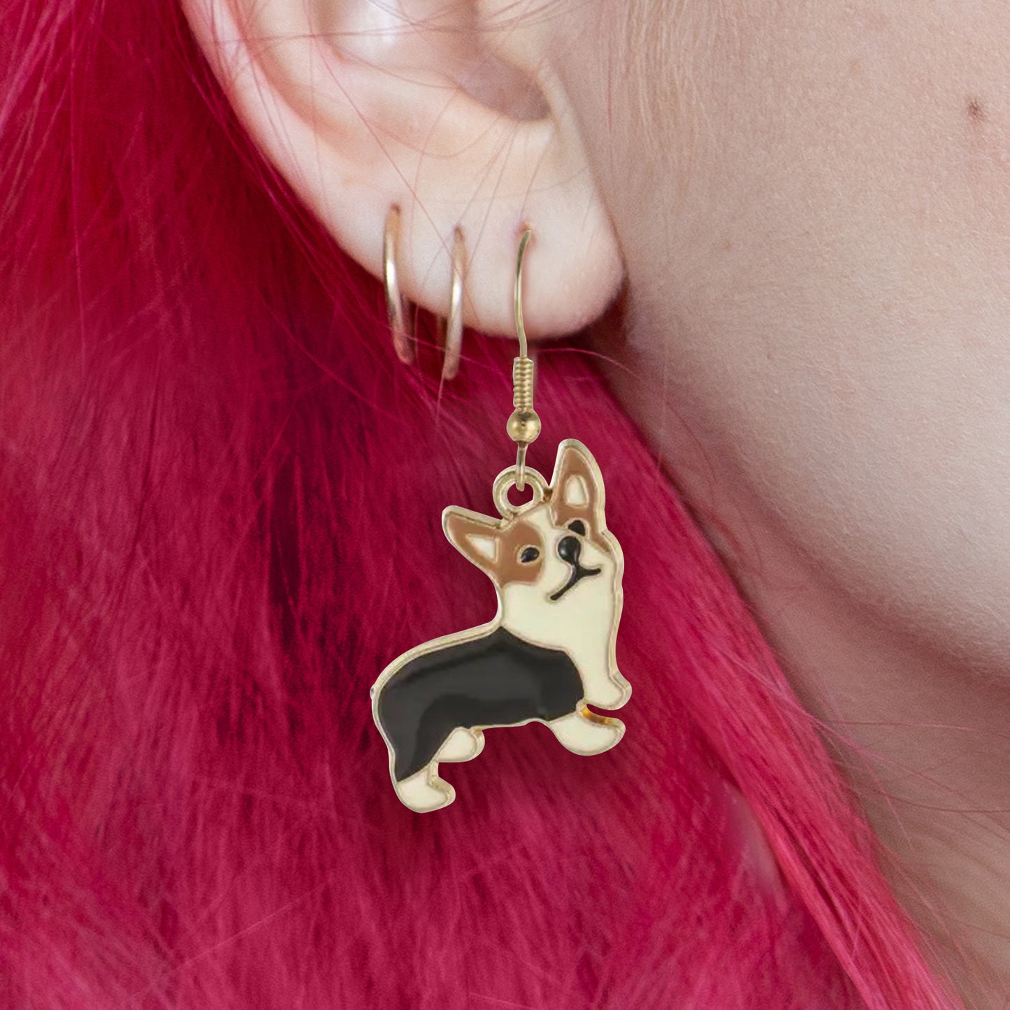Close up view of a model's ear. An earring shaped like a Welsh Corgi is hanging from her earlobe. Behind the earring is the model's bright pink hair.