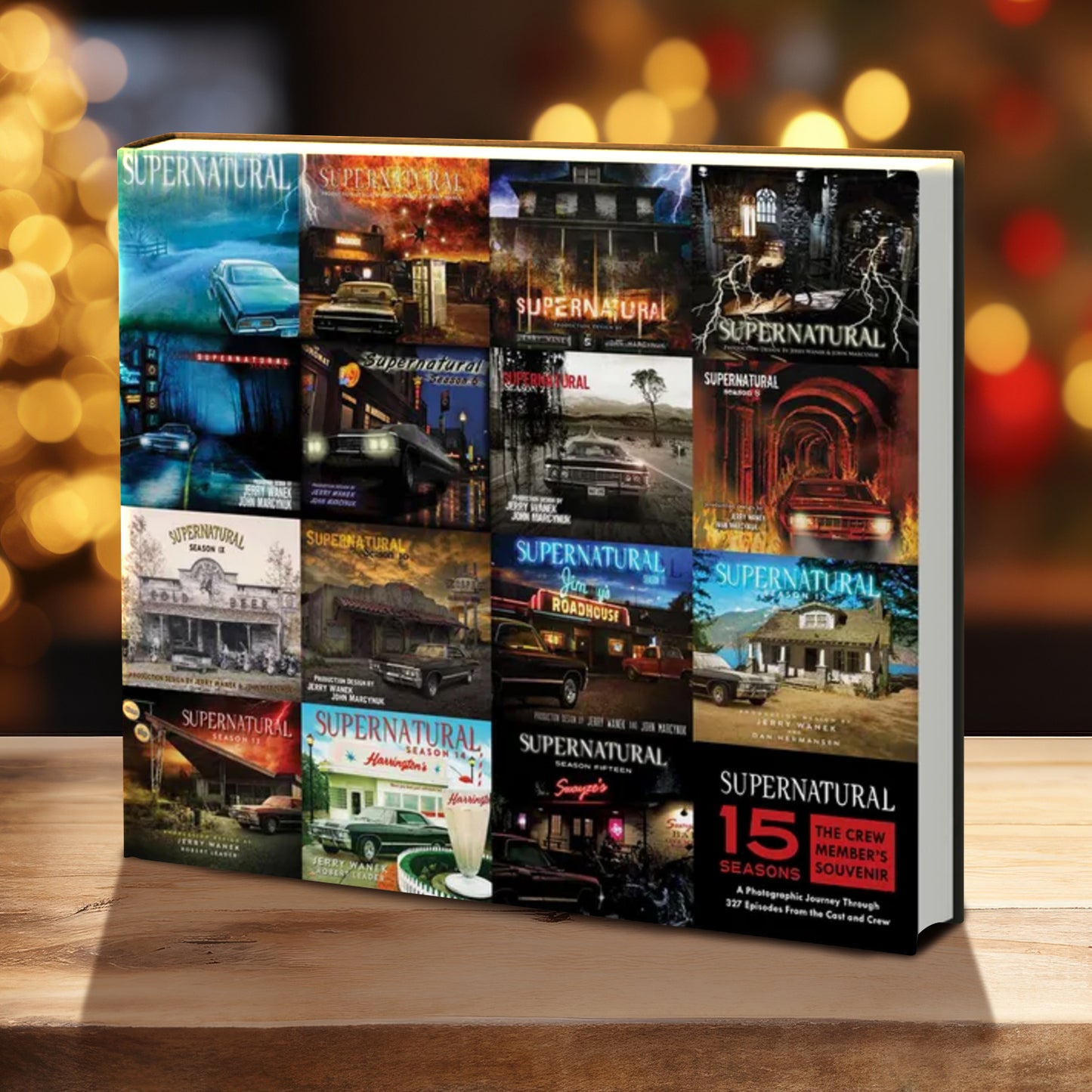 A book on a wood table, in front of twinkling Christmas lights. The book cover is made up of a a collage of images from15 seasons of the TV series Supernatuural. At the bottom right corner is a black square with white and red text saying "supernatural, 15 seasons: the crew member's souvenir. A photographic journey through 327 episodes from the cast and crew.