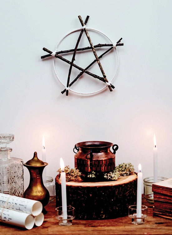 A pentagram fashioned from twigs hanging on a white background. Below the pentagram is a wooden table, with scrolls, candles, and jars. In the center is a small tree stump, with a copper pot sitting on top of it.