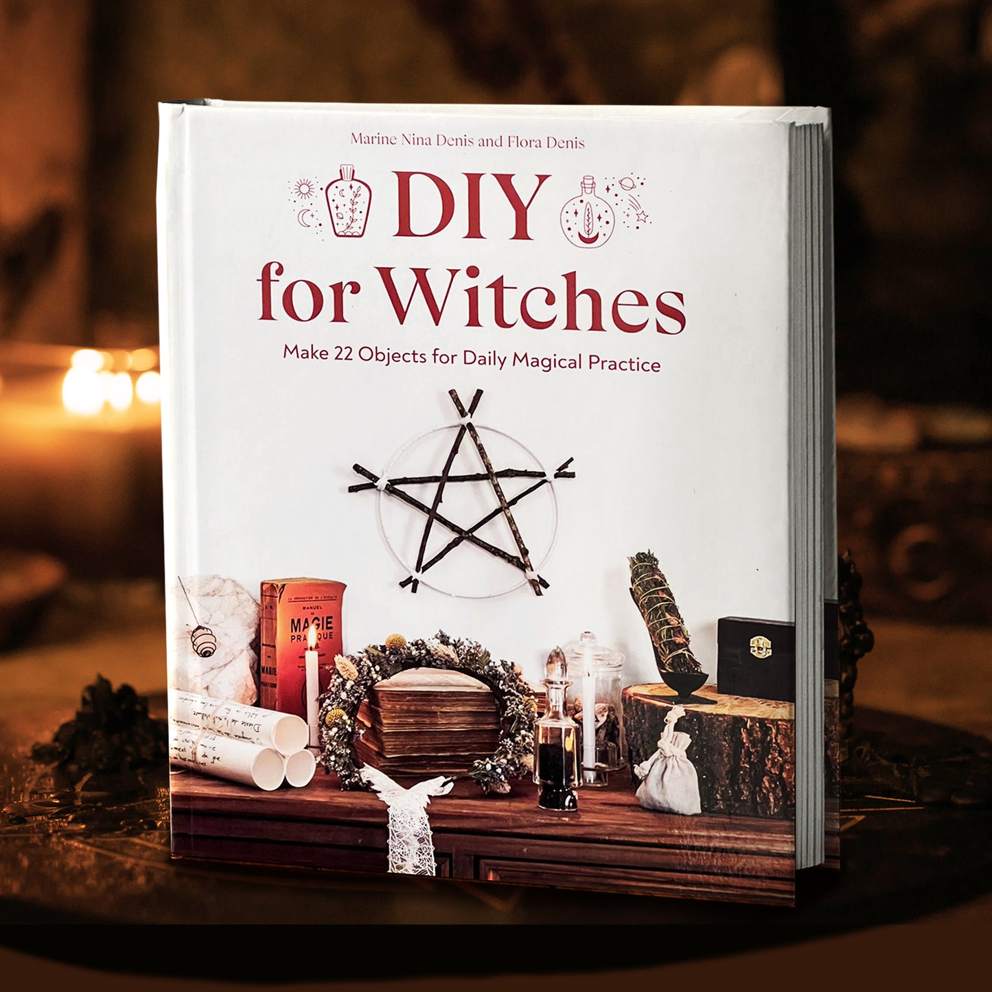 A white book on a circular tablet, with candles and spell ingredients in the background. On the cover is maroon text reading "DIY for witches." Under the text is a pentagram fashioned from twigs, hanging on a white background. Beneath the pentagram are various scrolls, books, candles, and other magical items, on a wooden cabinet.
