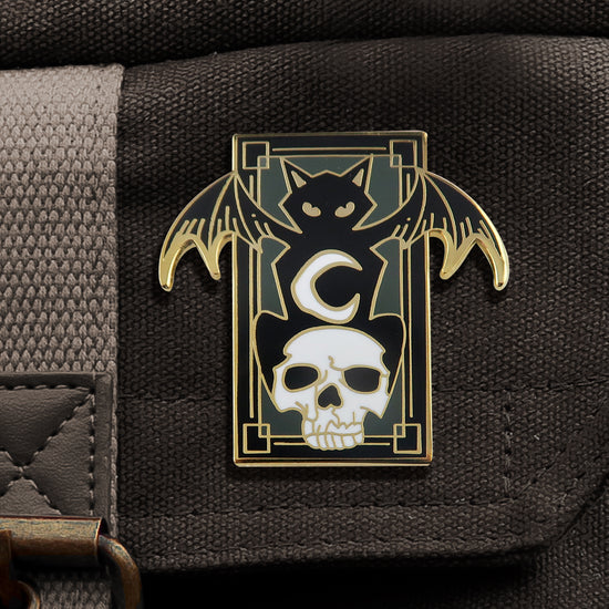 A rectangular, black enamel pin with gold edges, attached to a black canvas bag. On the front is a depiction of a black cat with bat wings, crouching on a white skull. On the cat's belly is a white crescent moon. Behind the cat is a green background.