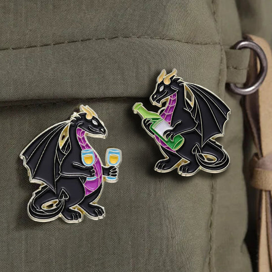 Two enamel pins side by side. Both pins depict black dragons with purple bellies. The left dragon is holding two wine glasses, the right dragon is holding a green wine bottle.