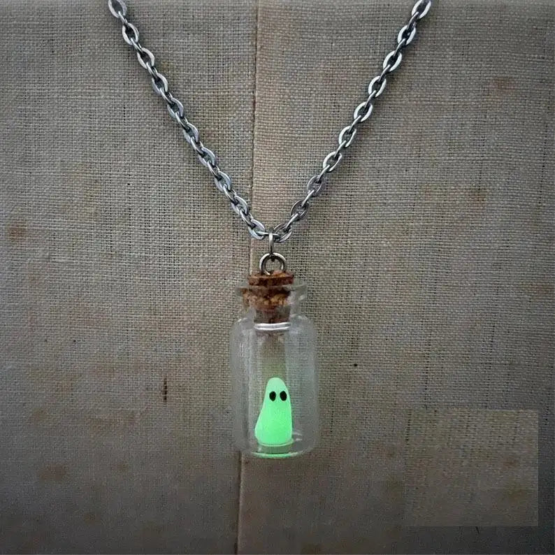 Close up view of a necklace hanging in front of a yellow and white canvas. The necklace is made of a tiny glass bottle with a cork stopper at the top. Inside the bottle is a tiny green "sheet" ghost with black eyes.