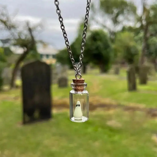 Close up view of a necklace suspended in the air, with a graveyard visible behind it. The necklace is made of a tiny glass bottle with a cork stopper at the top. Inside the bottle is a tiny green "sheet" ghost with black eyes.