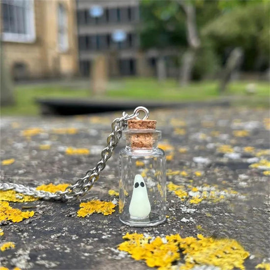 Close up view of a necklace standing on a concrete table, covered with white and yellow flower petals. The necklace is made of a tiny glass bottle with a cork stopper at the top. Inside the bottle is a tiny green "sheet" ghost with black eyes. Behind the necklace is a residential building with trees and grass around it.