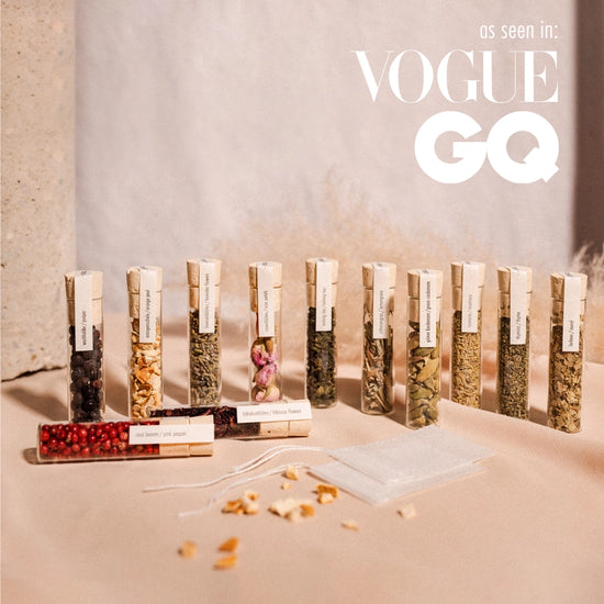 Twelve small glass vials, standing on a sand-colored floor. Inside each vial is a different botanical for cocktails, including hibiscus, lemongrass, orange peel, and others. Small, white cinch bags are next to the vials, used to infuse the botanicals into gine. At the top right corner is white text saying As seen in Vogue, GQ."