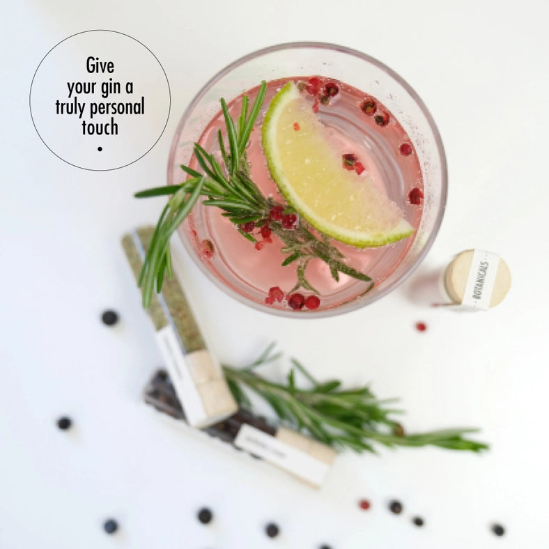 Top view of a cocktail glass filled with gin, with red peppercorns, thyme, and a lemon wedge inside. Next to the glass are small vilas of botanicals for cocktails. At the top left corner is a black circle with black text inside saying "give your gin a truly personal touch."
