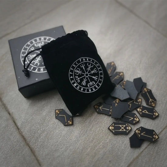 A small black velvet bag adorned with rune symbols, leaning against a black box with the same pattern. In front of the bag are scattered about 20 small woodcut runes with gold printing.
