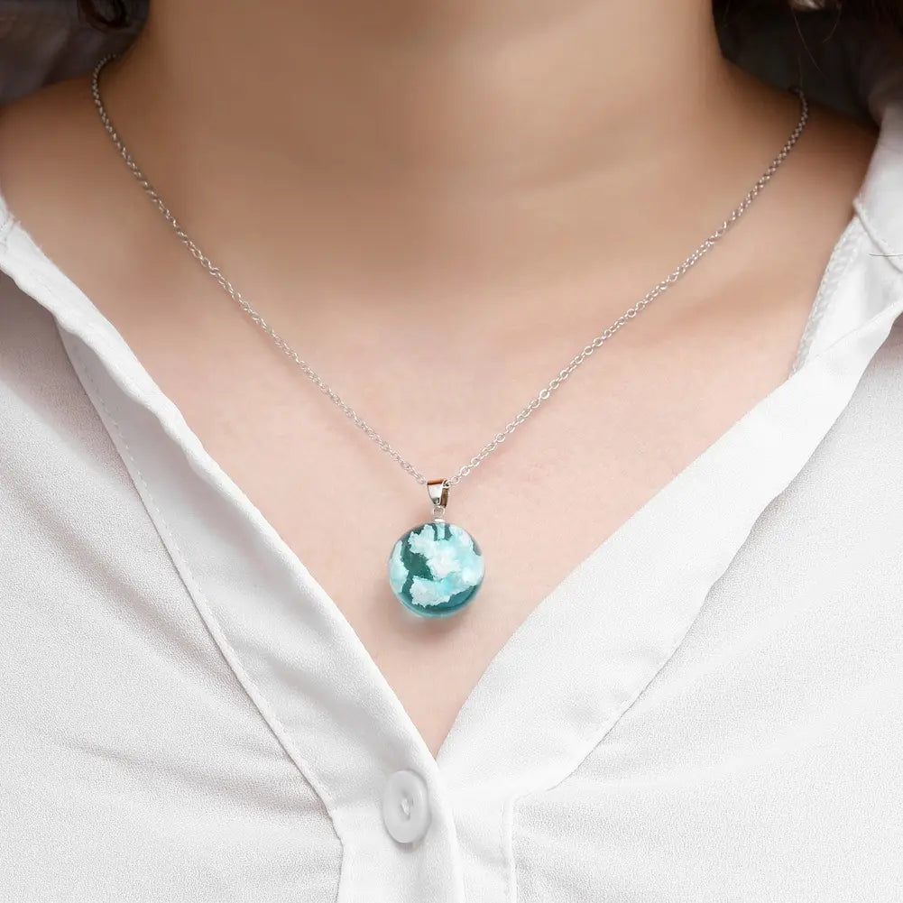 Close up of a model wearing a round, sky-blue pendant on a silver chain. The pendant is a sphere, with white clouds inside it.