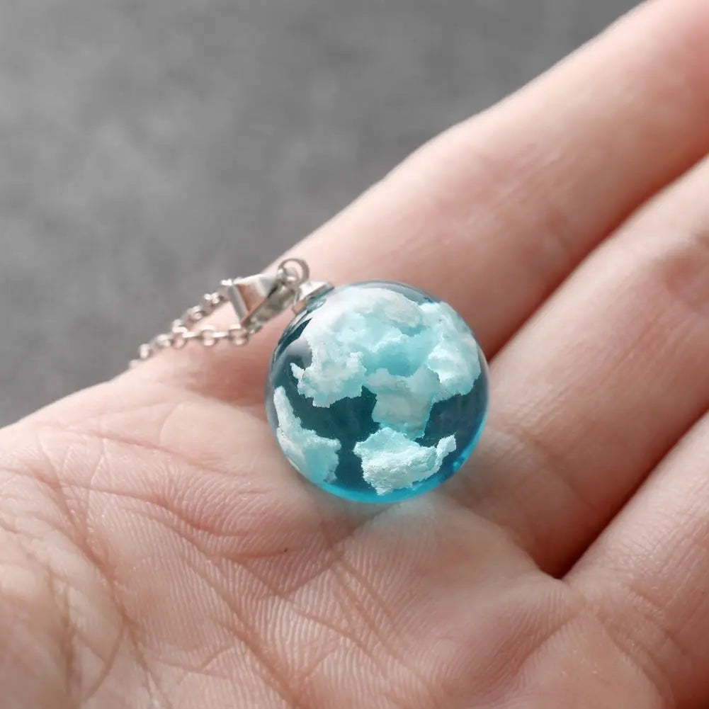 A round, sky-blue pendant on a model's hand. The pendant is a sphere, with white clouds inside it. A silver chain is attached at the top.
