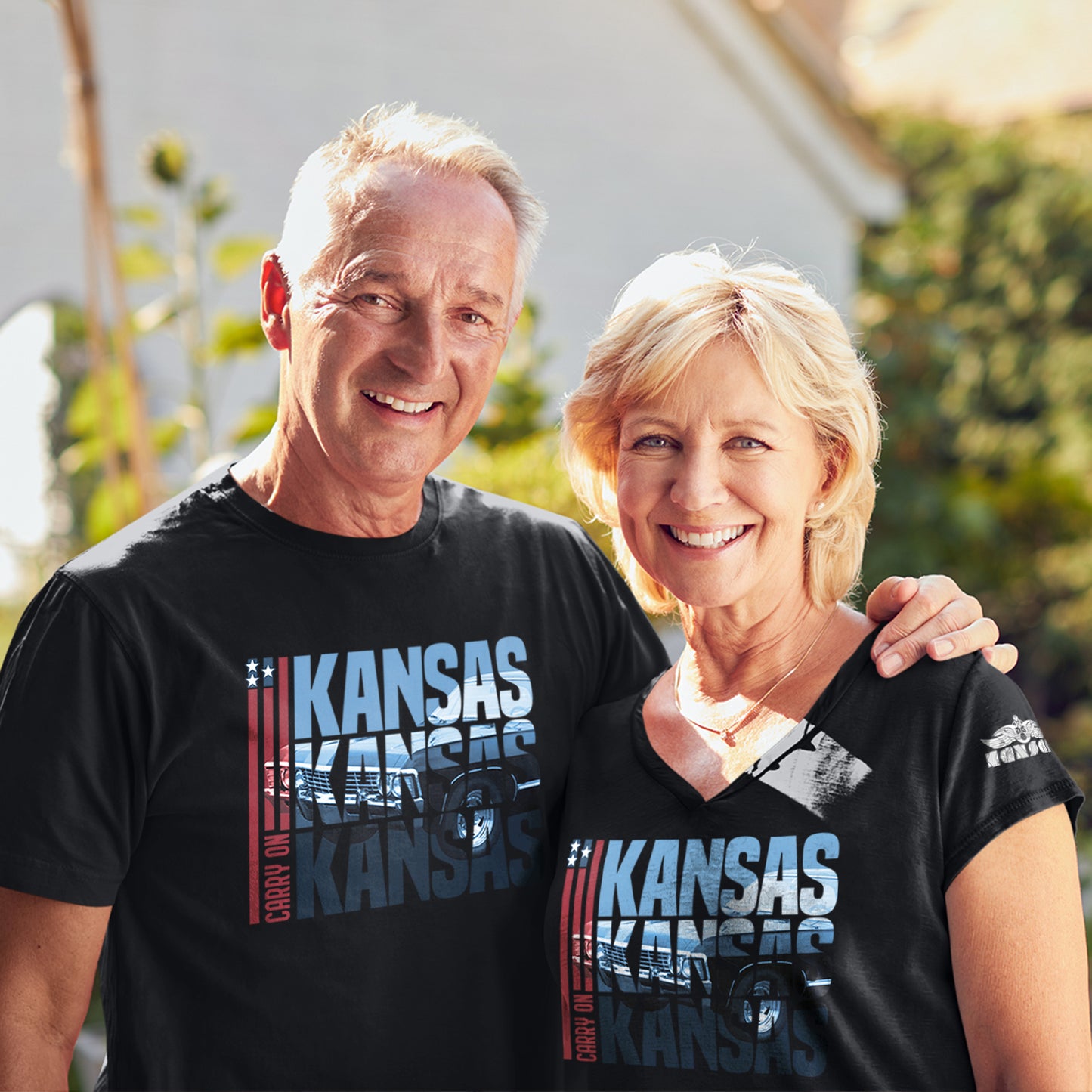 Two models side by side wearing black tee shirts. In the front of each shirt is blue text that says “Kansas Kansas Kansas,” with a black Chevy Impala superimposed on the text. On the left of the text are three red stripes with red text saying “carry on.”