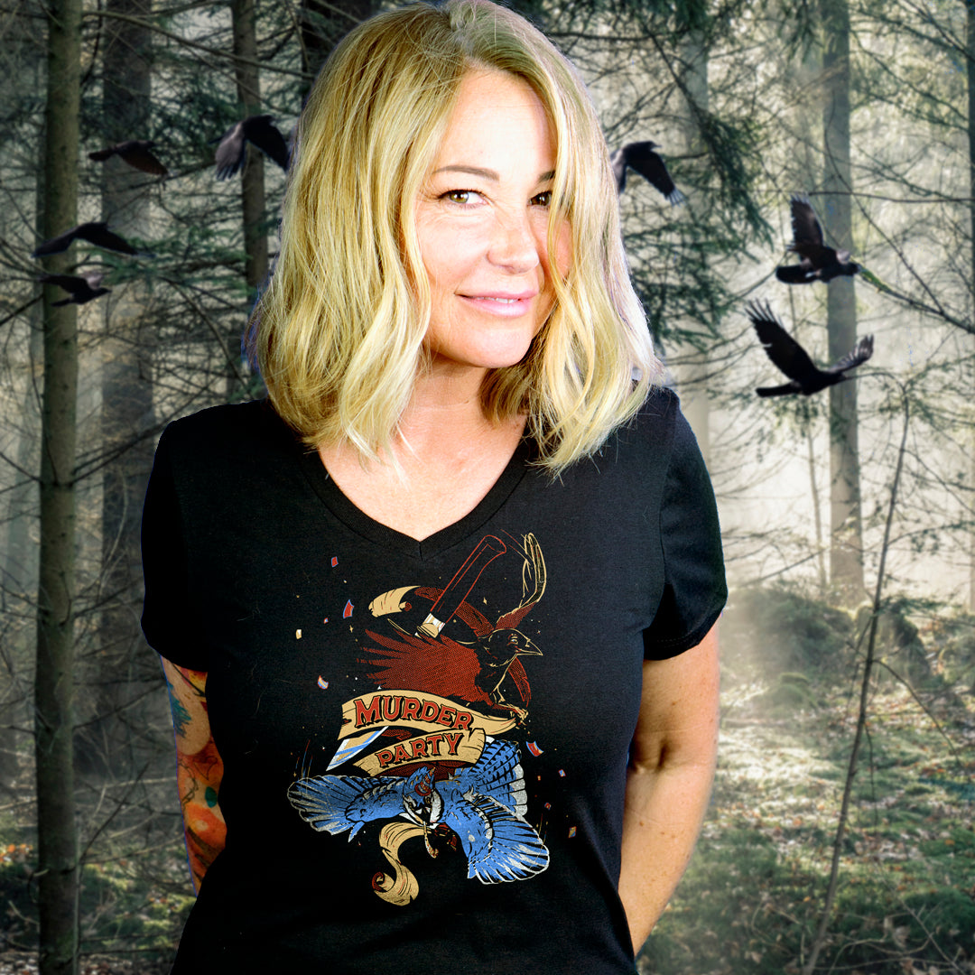 An image of the actress Kim Rhodes standing in front of a desolate forest. She is wearing a black T-shirt with a drawing of two crows on the front, one in red, the other in blue. At the center in red text is "murder party."