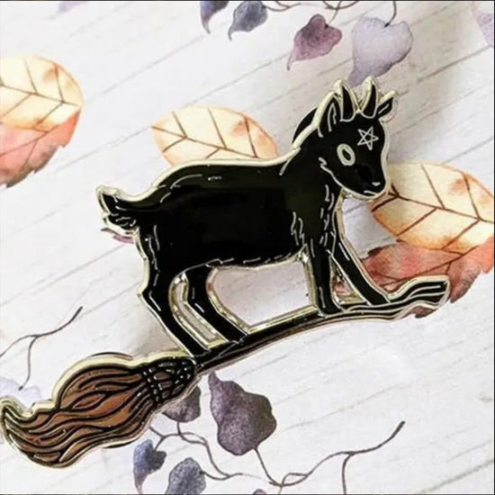 Close up of a black enamel pin in the shape of a baby goat standing on a broomstick. The goat has a five-pointed star drawn on its forehead. Behind the goat is a pattern of leaves in various colors against white wood.