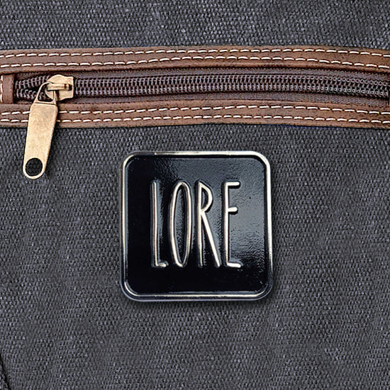 A silver enamel pin with a black rectangular front, which reads LORE in tall, skinny white font. The pin is affixed to a black canvas bag.