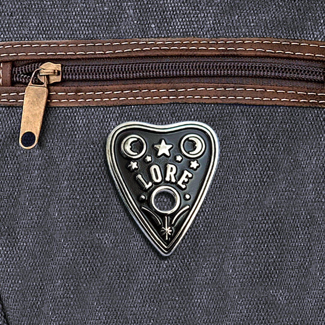 A silver enamel pin in a triangular shape resembling a OUIJA planchette with stars and two crescent moons, which reads LORE in tall, skinny white font. The pin is attached to a black canvas bag.