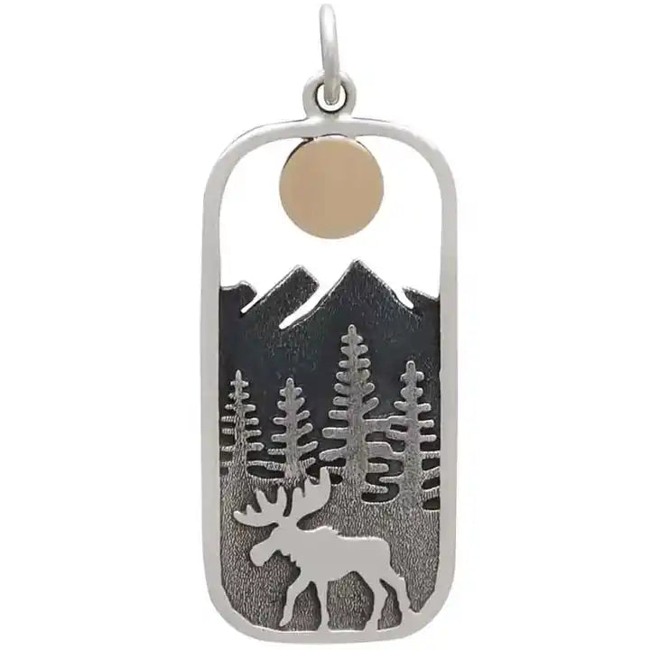 A rectangular pendant with rounded edges, against a white background. Inside the pendant is a depiction of a moose walking on front of pine trees, with mountains in the back, and a round golden sun at the top.