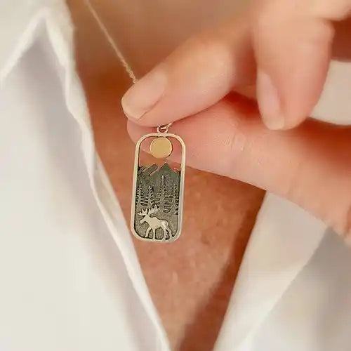 Close up of a model's hand holding a rectangular pendant with rounded edges, against the model's neck. Inside the pendant is a depiction of a moose walking on front of pine trees, with mountains in the back, and a round golden sun at the top. A stainless steel necklace chain is attached to the pendant.