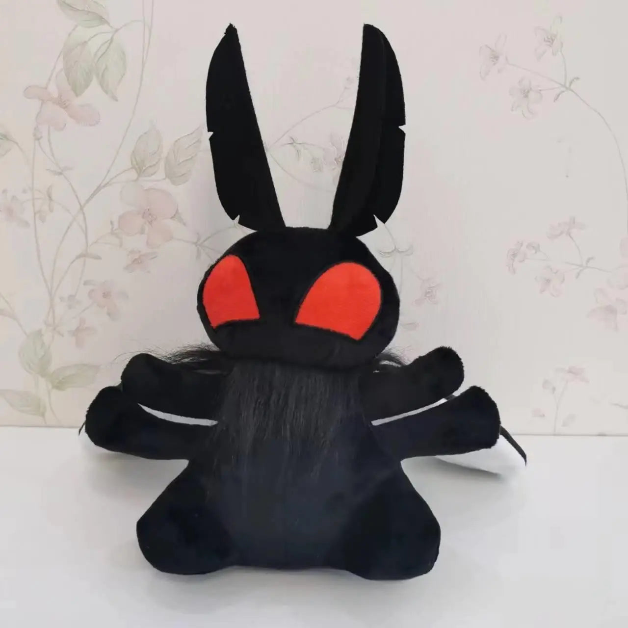 A black plushie shaped like the cryptid Mothman. The plushie has giant red eyes, feather-shaped antennae, and two rounded wings. Behind the plushie is wallpaper with a flower pattern.