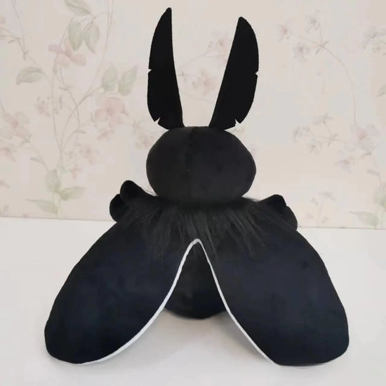 A black plushie shaped like the cryptid Mothman. The plushie has giant red eyes, feather-shaped antennae, and two rounded wings. Behind the plushie is wallpaper with a flower pattern.