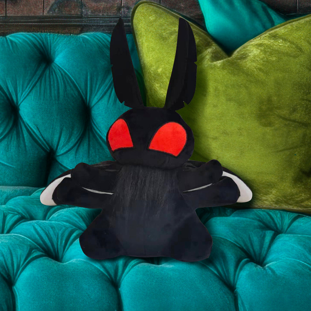 A black plushie shaped like the cryptid Mothman. The plushie has giant red eyes, feather-shaped antennae, and two rounded wings. The plushie is resting on a blue and green couch.