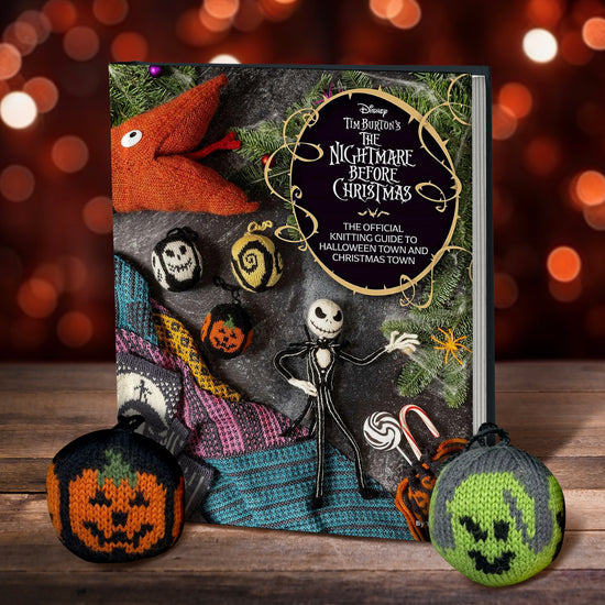 A book on a wood table. On the top right corner of the cover is a black oval, with white text saying "Tim Burton's The Nightmare before chrsitmas: the official knitting guide to halloween town and christmas town." On the cover are various knitting projects depicting characters from the film The nightmare before christmas. next to the book are knit halloween ornaments. In the background are red and orange lights.