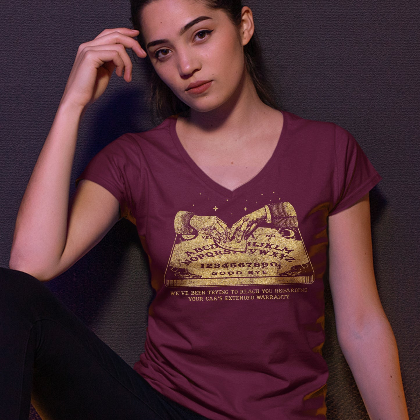 A female model wearing a maroon V-neck shirt, with a gold color image of two hands over a Ouija board. Under the board is gold text saying "We've been trying to reach you regarding your car's extended warranty." Behind the model is a dark purple background.