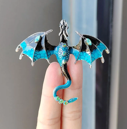 Close up view of a dragon-shaped brooch, sitting on a model's fingers.. The dragon is blue with silver accents, and has blue rhinestones inset in its tail and abdomen. The dragon's wings are spread out to the side, and its head is pointed straight out.