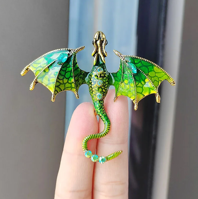 Close up view of a dragon-shaped brooch, sitting on a model's fingers.. The dragon is green with gold accents, and has green rhinestones inset in its tail and abdomen. The dragon's wings are spread out to the side, and its head is pointed straight out.
