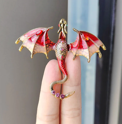 Close up view of a dragon-shaped brooch, sitting on a model's fingers.. The dragon is red with gold accents, and has red rhinestones inset in its tail and abdomen. The dragon's wings are spread out to the side, and its head is pointed straight out.