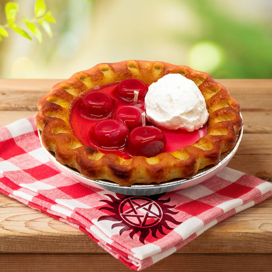 Close up view of a candle shaped like a cherry pie with whipped cream, sitting on a wooden table. Under the candle is a red and white checkered napkin with the anti-possession symbol on one corner.