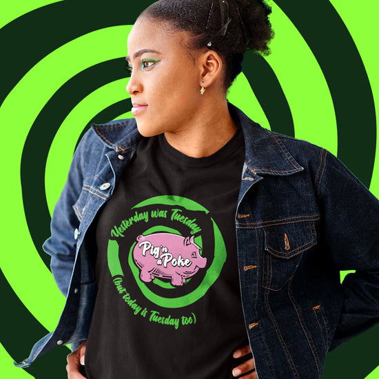 A female model wearing a black t-shirt under a dark denim jacket. The front of the tee has a bright-green-and-black swirl and within the swirl reads "Yesterday was Tuesday (but today is Tuesday too)". There is a pink pink in the middle and white font that reads "Pig n' a Poke'.