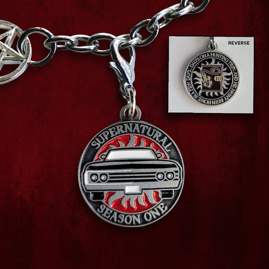 A brass coin charm with "Supernatural season one", a black Impala and an anti-possession symbol on one side and "Dad's on a hunting trip, and he hasn't been back in a few days" with a brown journal and amulet on the other. Behind the charm is a red wall.