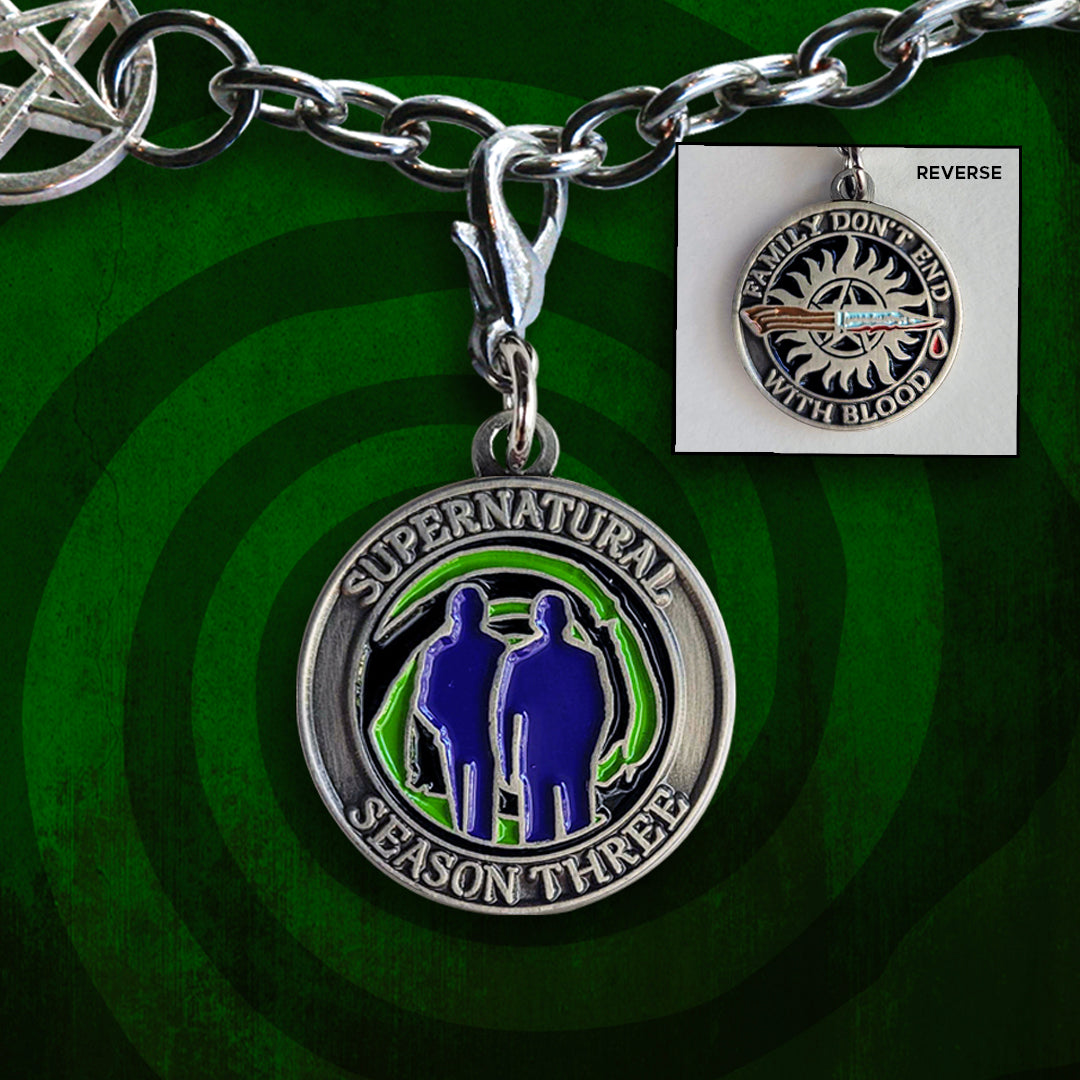 A brass coin charm with "Supernatural season three", a green "Mystery Spot" swirl, and 2 male sillhouettes on one side and "Family Don't End With Blood" with a knife and anti-possession symbol on the other. Behind the charm is a green wall with a dark green swirl