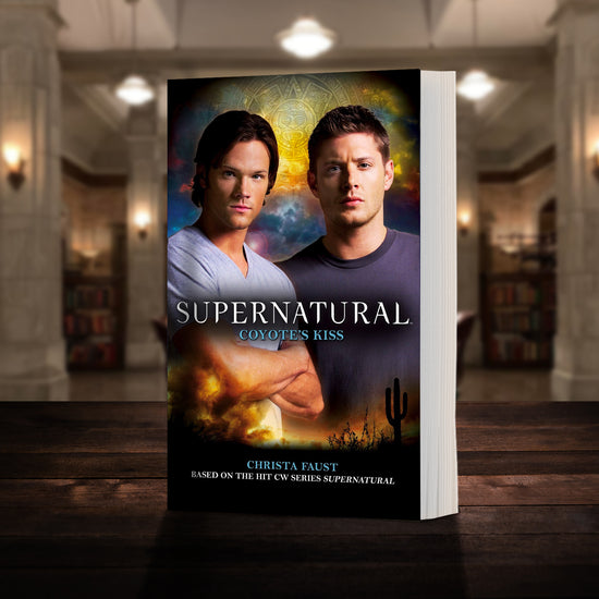A copy of the book "Supernatural: Coyote's Kiss" pictured in the "bunker" from Supernatural. The cover shows early-season Sam and Dean Winchester and reads "Supernatural Coyote's Kiss - Based on the hit CW series SUPERNATURAL by Christa Faust”