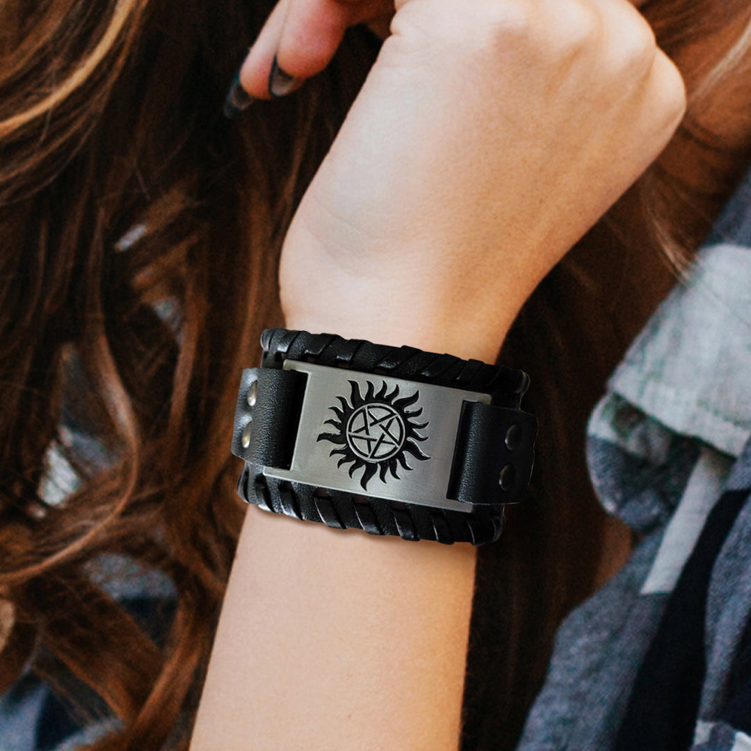 Close up view of a model's arm, with a black braided leather bracelet at the wrist. In the center of the bracelet is a metal plate with the anti-possession symbol etched onto it in black. Behind the arm is the model's hair against a blue plaid shirt.