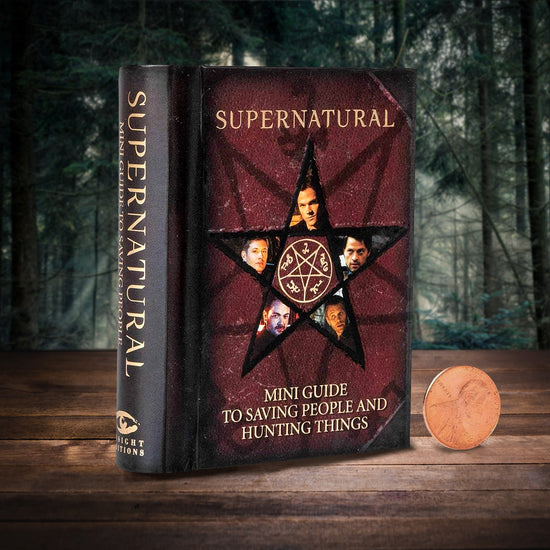An image of a dark red bookcover on a wood table, set against a forest background. In yellow text is the title "supernatural: mini guide to saving people and hunting things." In the middle of the cover is a five pointed start, with the anti-possession symbol in the star's center. Each point of the star features an image of a different character from the Supernatural TV series. Next to the book is a US penny, to show the size of the book.