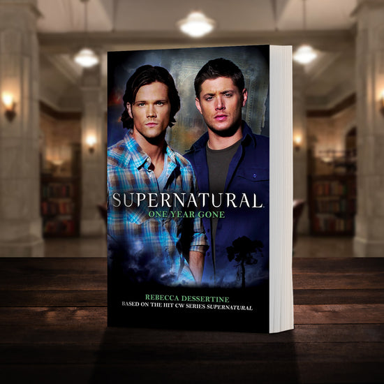 A copy of the book "Supernatural: One Year Gone" pictured in the "bunker" from Supernatural. The cover shows early-season Sam and Dean Winchester and reads "Supernatural One Year Gone - Based on the hit CW series SUPERNATURAL by Rebecca Desertine”
